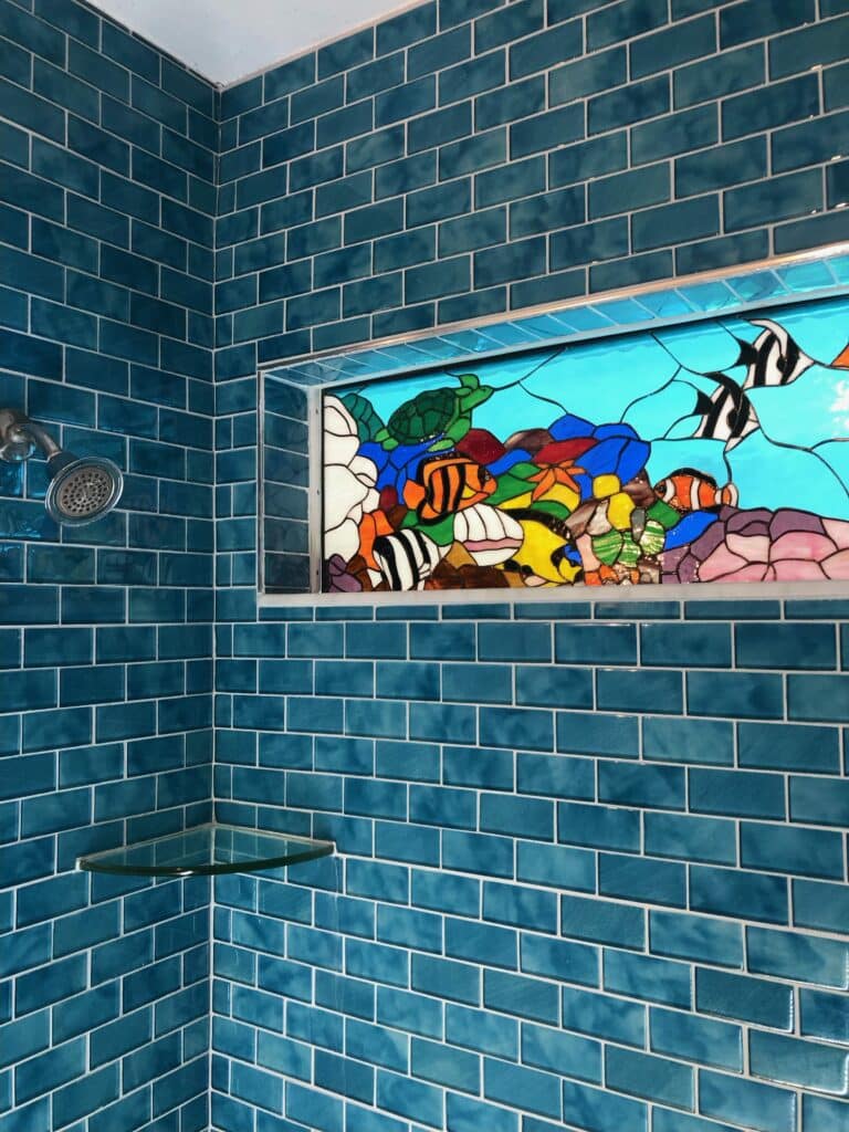 A vibrant stained glass panel illustrating an underwater coral reef teeming with colorful tropical fish, including clownfish, striped fish, and a green sea turtle, integrated into a rectangular shower niche. The shower walls are adorned with glossy teal blue subway tiles, and a chrome shower head is on the left.