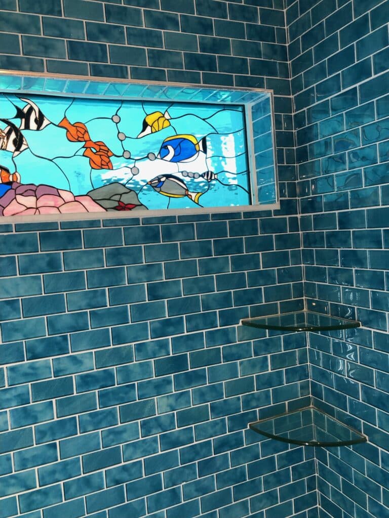 A rectangular leaded stained glass panel featuring a vibrant underwater scene. Colorful fish, including a black and white striped fish, an orange fish, a yellow fish, and a blue tang, swim amidst blue water and pink and grey coral. The panel is set into a wall covered with glossy teal subway tiles, with two clear glass corner shelves below it, suggesting a bathroom or shower environment.