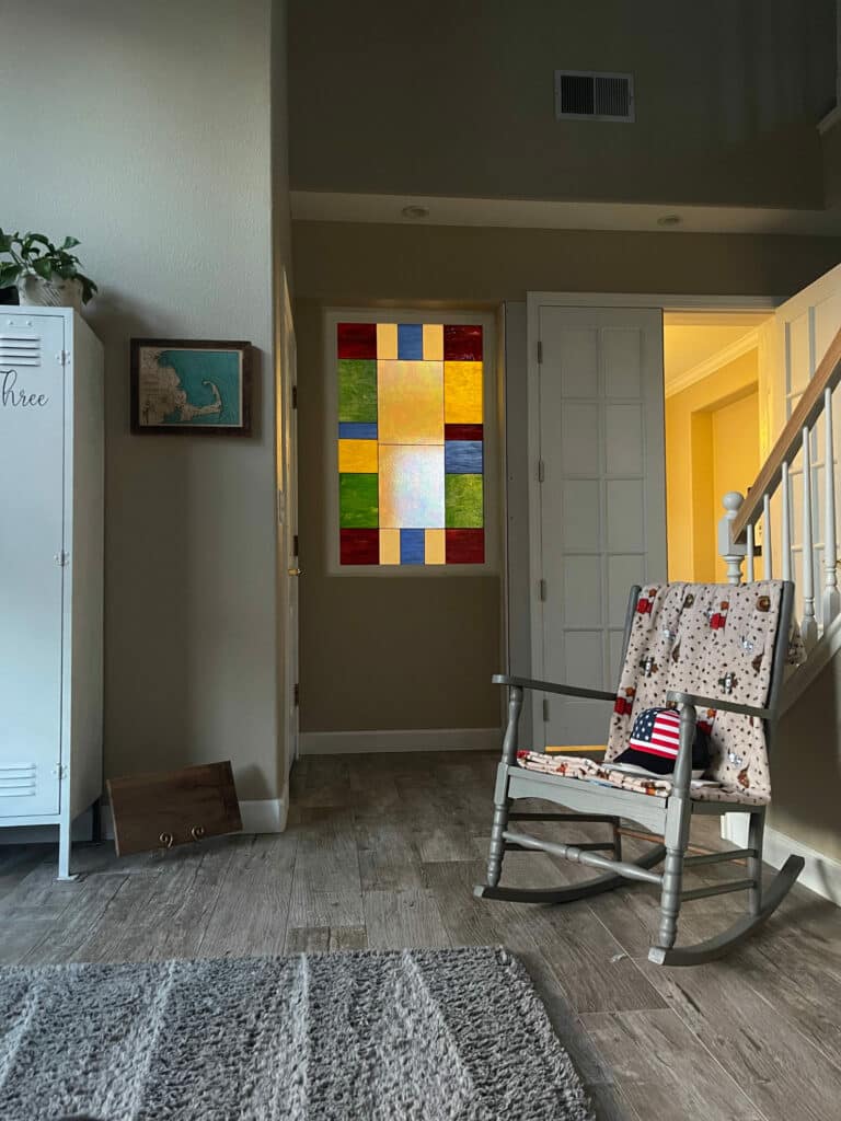 A rectangular stained glass window panel installed in a light beige wall. It features a modern geometric design with a large central square of clear, subtly textured glass, surrounded by smaller squares and rectangles of vibrant red, green, blue, and yellow stained glass.