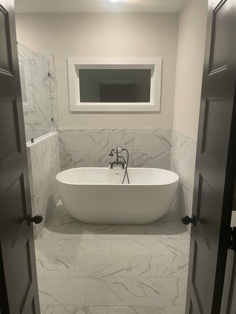 A custom rectangular stained glass privacy panel featuring clear beveled and textured glass, installed in a modern bathroom window above a freestanding white bathtub with dark fixtures, white marble-look tile walls, and floors.