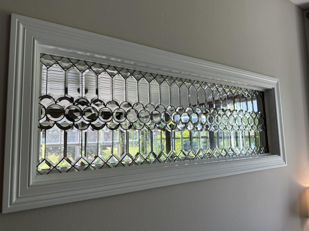 A rectangular clear beveled glass transom window panel with a white frame, featuring a central horizontal band of shimmering circular bevels and surrounding geometric diamond and elongated hexagonal shapes, allowing natural light through.