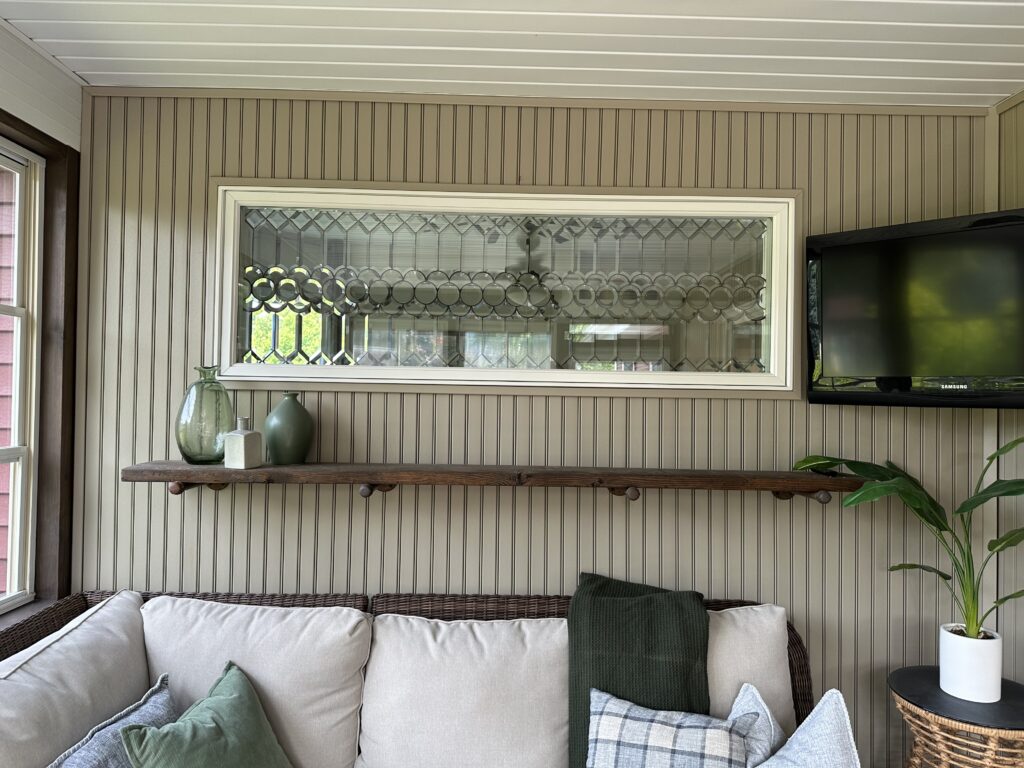 A horizontal leaded stained glass window panel with a clear, textured, and beveled geometric design. The top section features diamond and rectangular shapes, while the bottom section has a distinctive border of clear, round, scallop-like glass pieces. The panel is set in a white frame, providing privacy above a wooden shelf and wicker couch in a room with vertical slat walls.