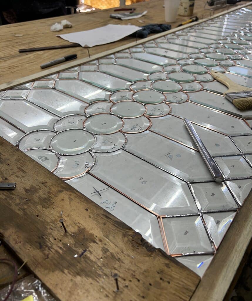 Close-up of a clear beveled stained glass panel being assembled on a wooden workbench. The intricate design features circular beveled glass pieces forming floral patterns, surrounded by larger geometric beveled and clear textured glass sections. Copper foil wraps the edges of the individual glass pieces, with some pieces numbered. A metal tool and a brush rest on the panel, showcasing the Tiffany method in progress.