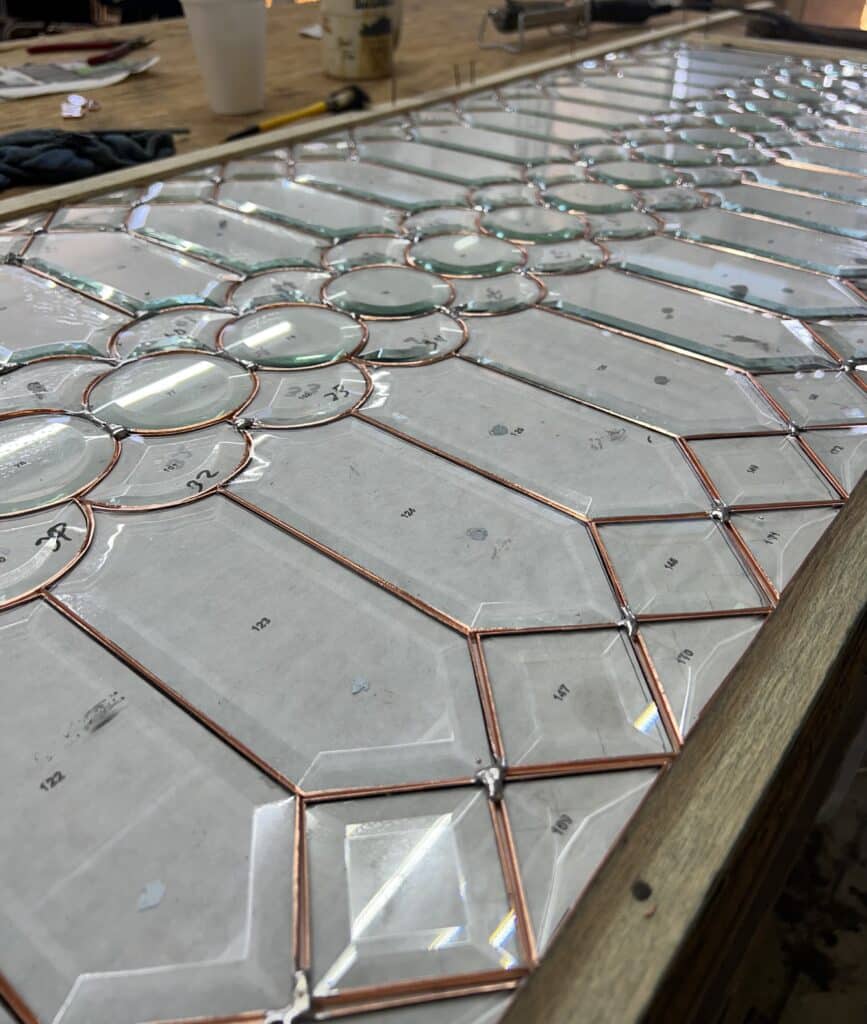 A close-up, angled view of a custom clear beveled stained glass panel laid flat on a workbench during its construction. Individual pieces of transparent glass, including circular jewels, square, rectangular, and octagonal bevels, are meticulously outlined with copper foil. Some glass pieces have handwritten numbers indicating their placement. Partial solder points connect the copper-foiled edges, showcasing the Tiffany method. A wooden jig holds the panel's edges, with workshop tools and a white cup blurred in the background.