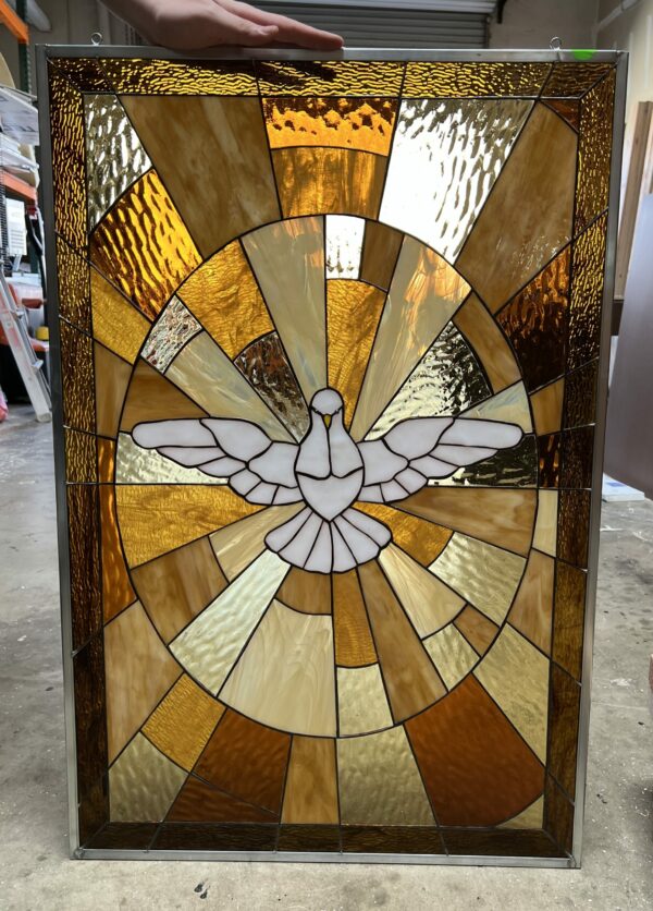 A rectangular stained glass panel depicting a white dove with outstretched wings, symbolizing the Holy Spirit, at its center. The dove is surrounded by a radiating sunburst pattern of textured art glass in shades of amber, golden yellow, and warm brown, all joined by lead came and framed in metal.