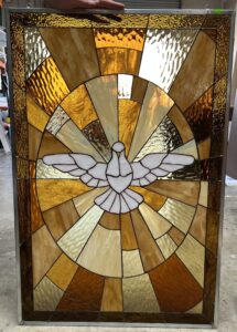 A rectangular stained glass panel depicting a white dove with outstretched wings, symbolizing the Holy Spirit, at its center. The dove is surrounded by a radiating sunburst pattern of textured art glass in shades of amber, golden yellow, and warm brown, all joined by lead came and framed in metal.