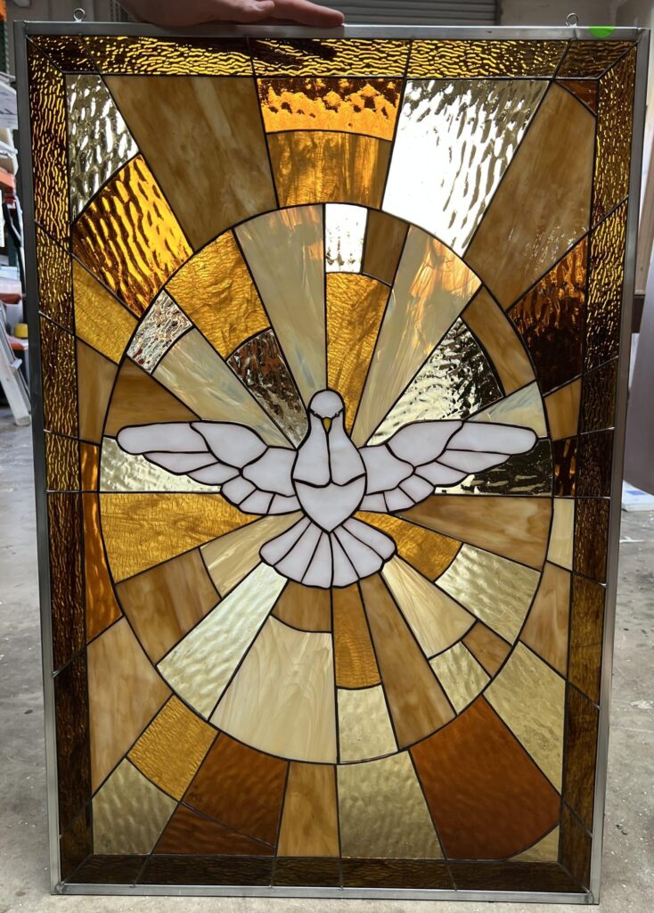 A rectangular stained glass panel depicting a white dove with outstretched wings, symbolizing the Holy Spirit, at its center. The dove is surrounded by a radiating sunburst pattern of textured art glass in shades of amber, golden yellow, and warm brown, all joined by lead came and framed in metal.