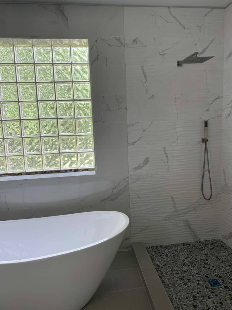 A contemporary bathroom featuring a large rectangular glass block window above a white freestanding bathtub. The walls are tiled with white marble-look ceramic tiles and a textured wavy white tile in the shower area, which includes a rain showerhead and a handheld shower. The shower floor is made of dark pebble mosaic tiles. Lush green foliage is visible through the glass block window, indicating natural light.