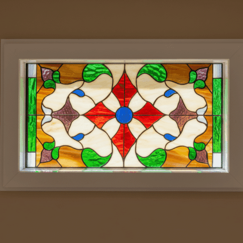 A rectangular, insulated vinyl window with an ornate Victorian stained glass panel inside. The symmetrical design features a central red star with a blue jewel, surrounded by intricate scrollwork in green, amber, and brown glass.