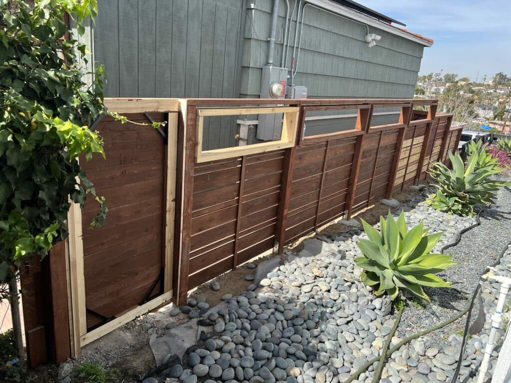 A newly constructed modern horizontal slat fence in dark stained wood with light wood framing for open rectangular inserts at the top. The fence runs along a landscaped area with river rocks and succulent agave plants, beside a house with grey siding.