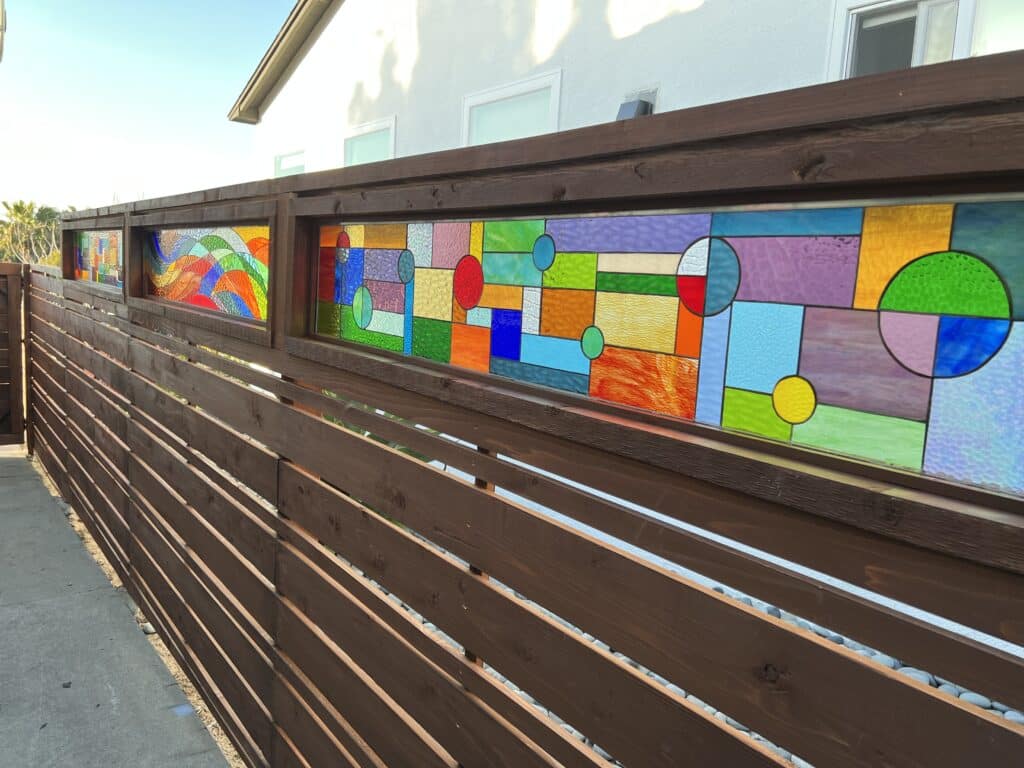 A long, dark stained wooden fence featuring three integrated rectangular stained glass panels under bright sunlight. The panels display vibrant, abstract designs: the leftmost panel shows colorful wavy lines, while the two rightmost panels feature a mosaic of geometric rectangles and circles in various colors and textures like red, blue, green, yellow, orange, and purple. The ground beneath the fence is covered in white pebbles, and a house is visible in the background.