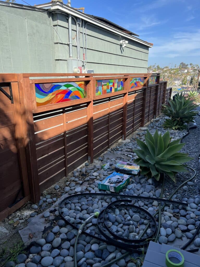 A series of dark brown horizontal slat wooden fence panels, each topped with a rectangular stained glass insert. The two outer stained glass panels feature flowing, abstract designs in vibrant shades of blue, green, orange, and red. The central stained glass panel displays a geometric mosaic pattern with multi-colored square and rectangular pieces. The fence is set in an outdoor area with grey river stones and large agave plants under a blue sky.