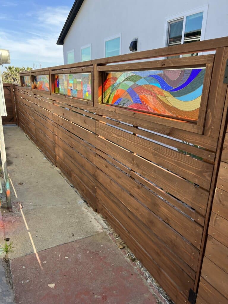A long, modern horizontal-slat wooden privacy fence featuring multiple rectangular stained glass art panels. The panels display vibrant, abstract designs, including one with flowing wave-like patterns in blues, greens, oranges, and reds, and another with a mosaic of multicolored, textured glass pieces. The fence borders a concrete path with a white house and blue sky in the background.