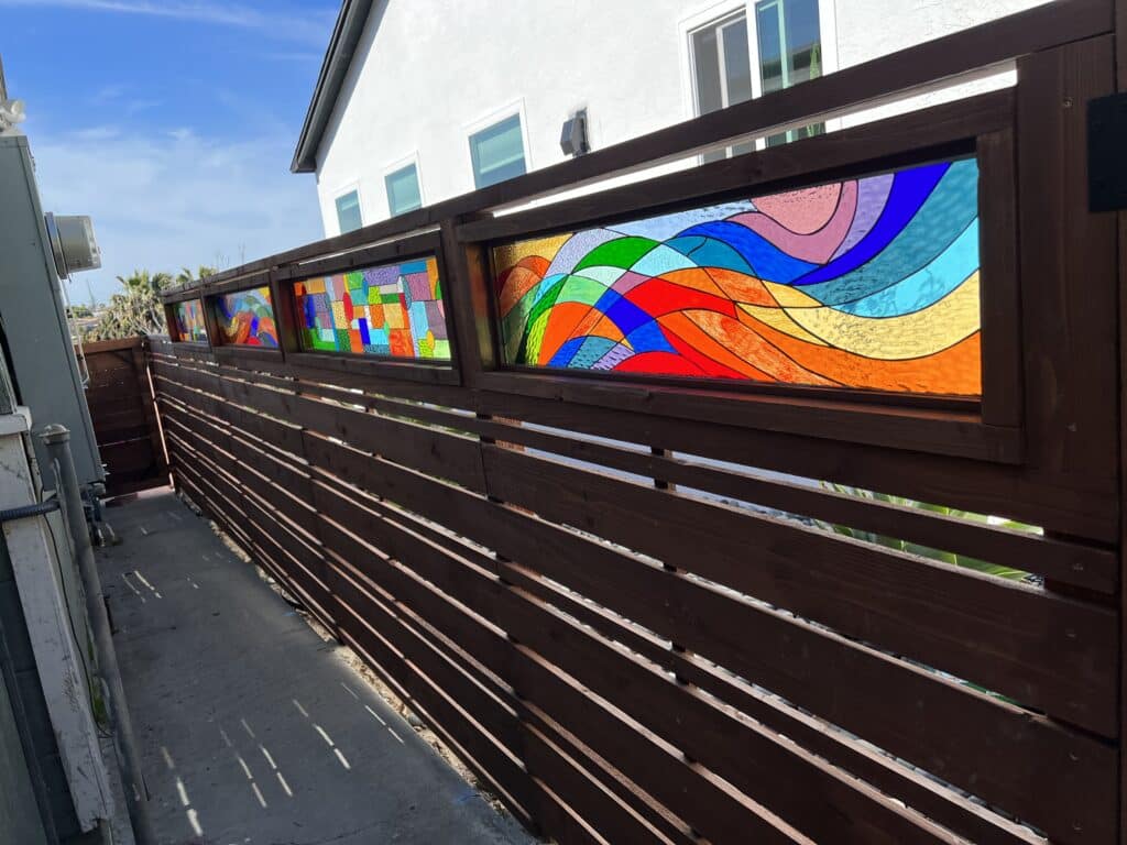 A series of rectangular, brightly colored abstract stained glass panels integrated into the top section of a dark brown wooden slat fence. The rightmost panel features a flowing, wavy design with sections of red, orange, yellow, green, blue, and purple glass. The central panel displays a more geometric, mosaic-like pattern of rectangles, squares, and circles in various vivid hues. The panels are set in an outdoor scene with a concrete path in the foreground, and a white house with windows and a blue sky in the background.