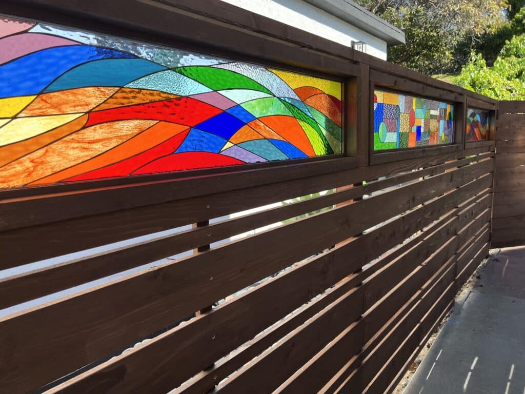 A dark brown wooden slatted fence featuring two integrated rectangular stained glass panels. The larger panel on the left displays an abstract wavy design in vibrant red, orange, yellow, green, and blue. The smaller panel on the right shows a geometric, mosaic-like abstract pattern with various colored glass textures.