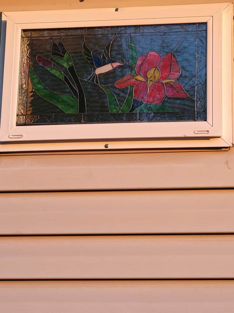 A rectangular leaded stained glass window, set within a white vinyl frame on beige house siding, depicting a vibrant hummingbird with a white belly and pink throat approaching a large red iris flower with a yellow center, surrounded by varied green leaves against a dark blue textured background.