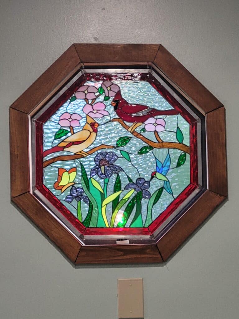 An octagonal stained glass panel in a dark wooden frame, depicting a vibrant nature scene with a red male cardinal and a yellow female cardinal perched on flowering branches, a blue and green hummingbird, a yellow butterfly, and purple irises, all against a textured blue sky.