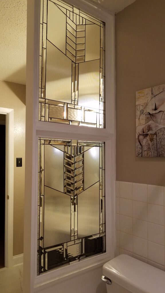 Two vertically stacked rectangular leaded glass panels, serving as an interior room divider. The panels feature a complex geometric design, blending clear, reeded, stippled, and heavily beveled glass pieces to create privacy and diffuse light in a Prairie or Art Deco style.