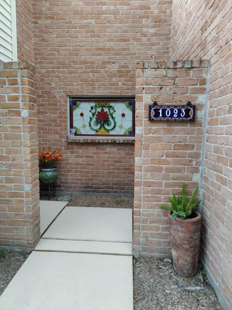 A rectangular stained glass panel featuring a vibrant red central flower with green scrolling foliage and circular blue, yellow, and red glass accents, set against a clear textured glass background. The panel is integrated into a rustic red brick wall, serving as a window in an outdoor entryway. To its right, a decorative blue and white ceramic tile house number plaque displays '1023'. The entryway includes light-colored concrete pavers, a terracotta planter with a green plant, and a green ceramic planter with orange flowers.