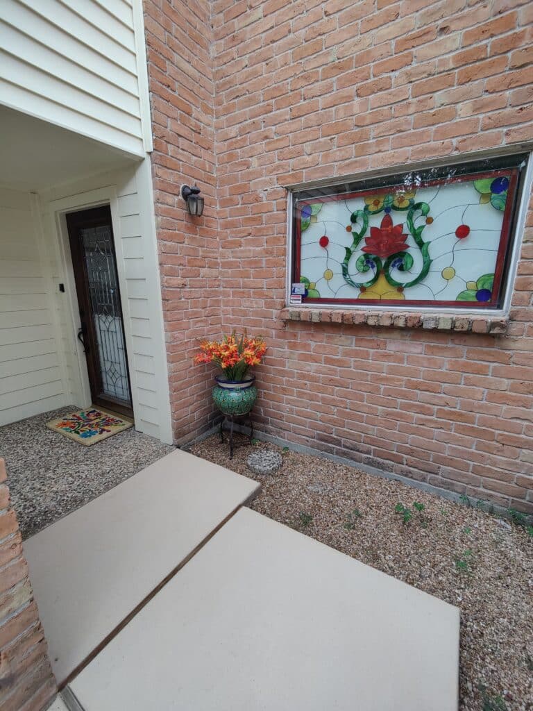 A rectangular exterior stained glass window featuring a vibrant red lotus flower design with green scrolling vines, clear textured glass, and colorful circular accents, set into a red brick wall next to a house entrance.