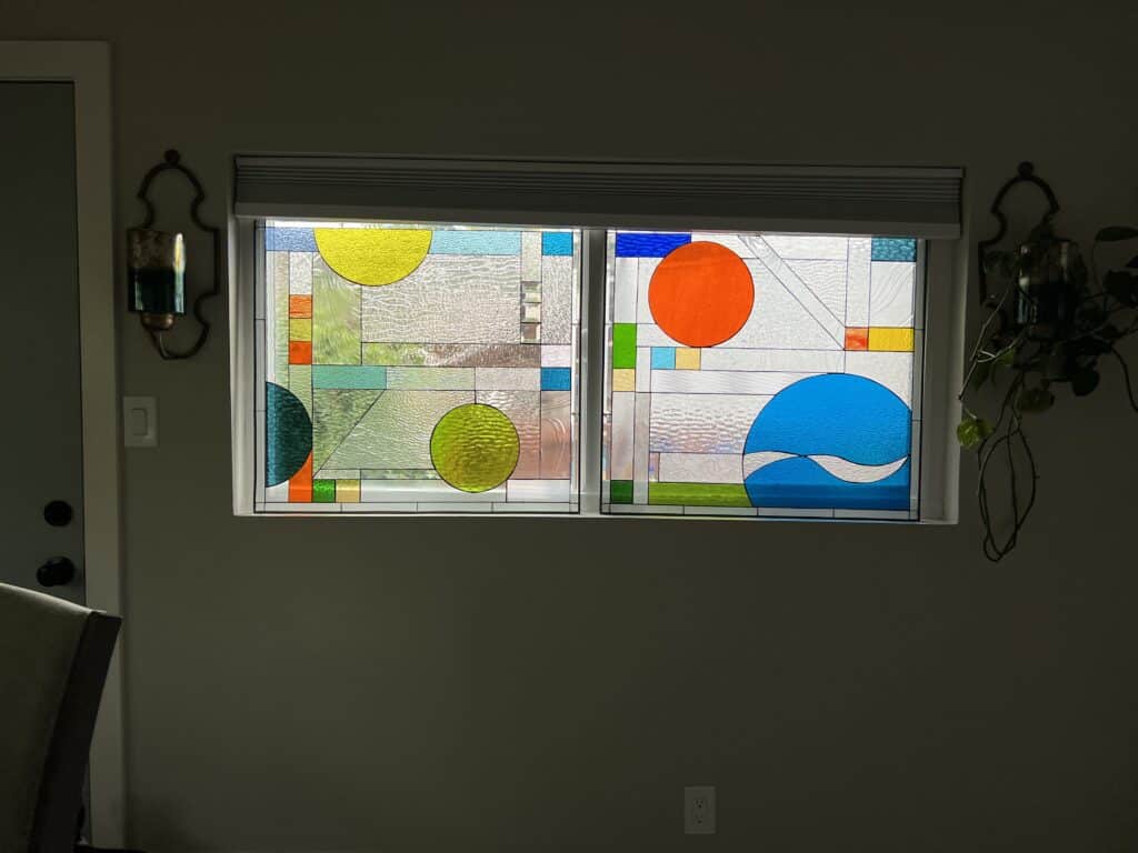 A rectangular stained glass window with two panels, each showcasing a modern abstract design. The panels feature large circular elements in yellow, orange, and blue, complemented by geometric clear textured glass sections and subtle wavy patterns. Additional accent colors are green, red, and light blue.