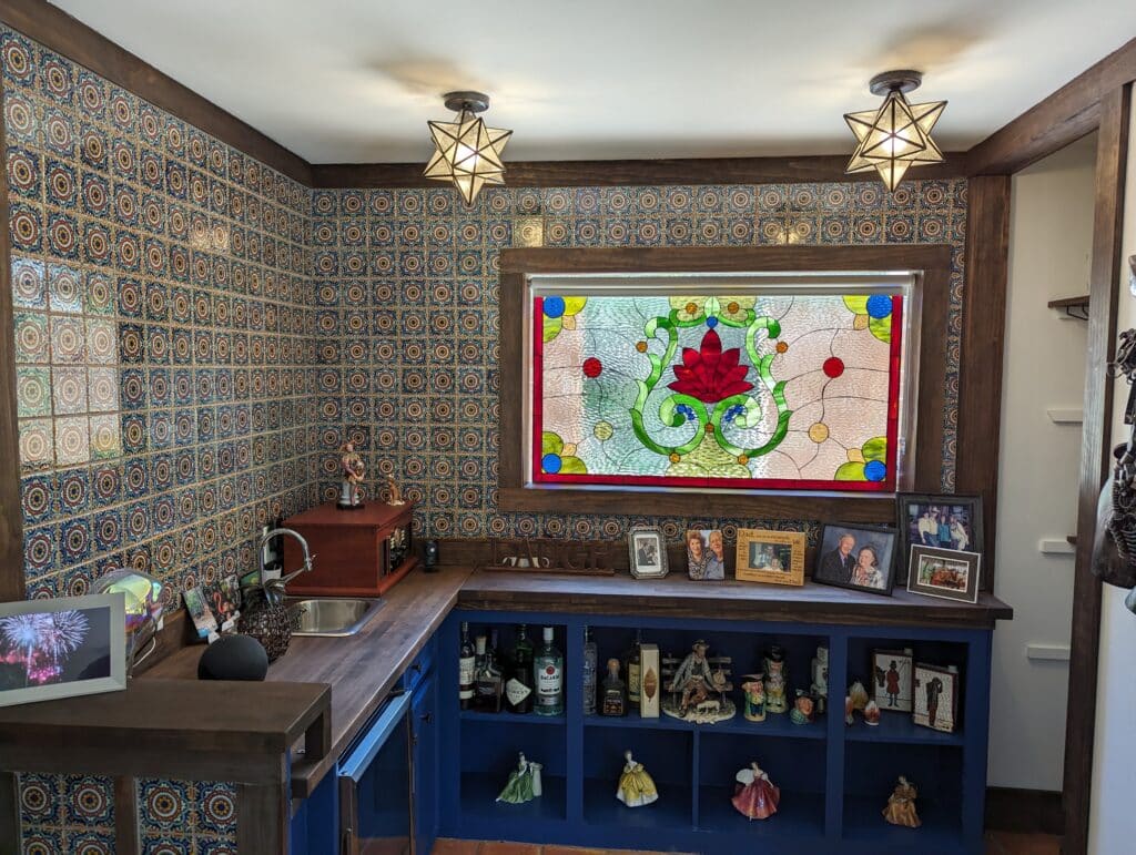 A rectangular stained glass window panel featuring a vibrant red lotus flower at its center, surrounded by intricate green swirling vines and leaves, set against clear textured glass with blue and amber circular accents, all framed by a bold red border. It is installed above a home bar with colorful patterned tiles.