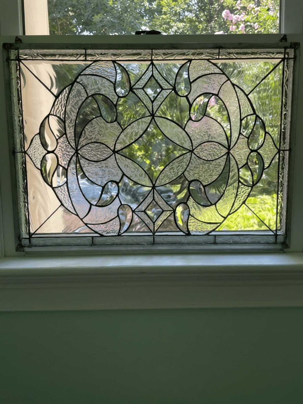 A rectangular, leaded stained glass panel with an ornate, symmetrical scrolling pattern made from clear textured and beveled glass, featuring curved elements and diamond accents, installed in a window.