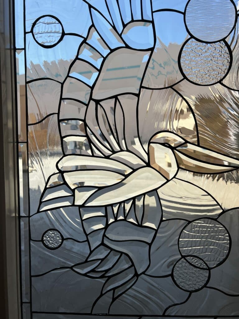 A leaded stained glass panel depicting an abstract, dynamic pelican or crane figure. The artwork uses various clear beveled glass pieces for the bird's body, wings, and long beak, creating faceted reflections. The background is composed of clear textured glass, including wavy and stippled patterns, with some light blue clear glass sections, and includes circular textured glass elements. The entire piece is framed by thin lead lines.