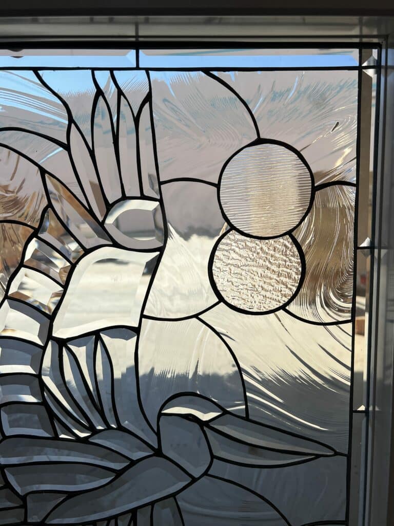 A rectangular stained glass panel with black lead lines, showcasing an abstract design made from various clear, textured, and beveled glass pieces. The design features dynamic flowing lines, fan-like patterns, and two circular inserts with distinct reeded and crinkled textures, all reflecting and refracting natural light.