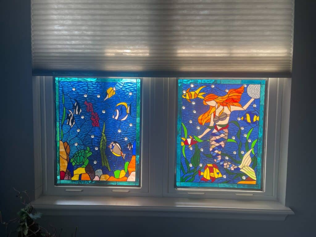 Two rectangular stained glass window panels installed side-by-side. The left panel depicts a vibrant underwater scene with blue water, colorful fish, a green sea turtle, and multi-colored coral. The right panel features a mermaid with long red hair and a blue-scaled tail, surrounded by fish and green seaweed. Both panels have textured blue backgrounds with clear circular jewels resembling bubbles and are framed with a turquoise glass border.