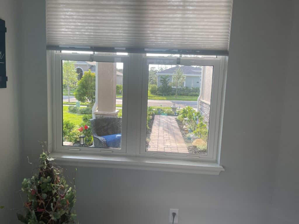 A rectangular window with a white frame and a partially lowered light grey honeycomb blind, looking out onto a sunny residential street with a brick paver walkway leading to a front porch, green landscaping, and houses.