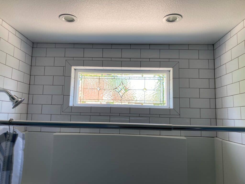 A rectangular, white-framed, leaded stained glass window installed high in a white subway tiled shower wall. The window features clear textured glass with a central geometric diamond motif accented by small light green and clear bevels, providing privacy while allowing natural light.