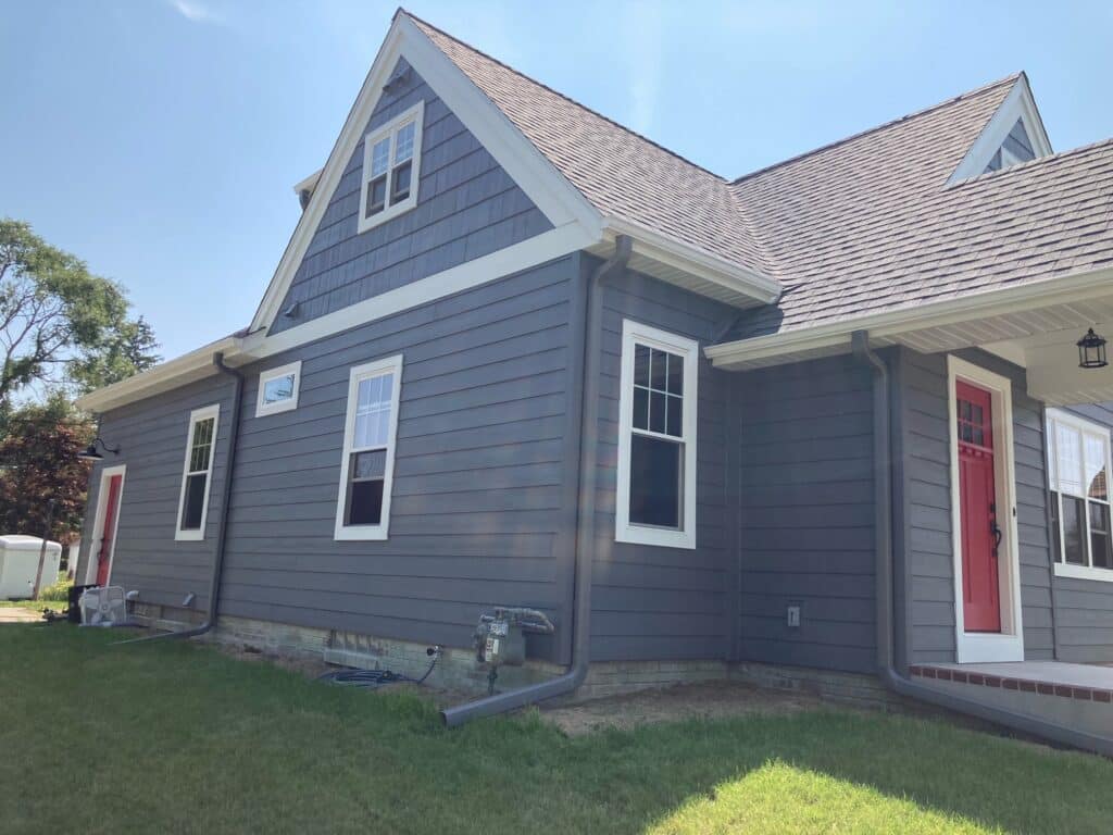 Exterior view of a modern farmhouse-style house with dark gray horizontal siding, crisp white trim, and two vibrant red entry doors. The home features multiple white-framed windows, a gabled roofline with a window, and brown shingle roofing under a clear blue sky, presenting an ideal setting for custom stained glass panels or transoms.