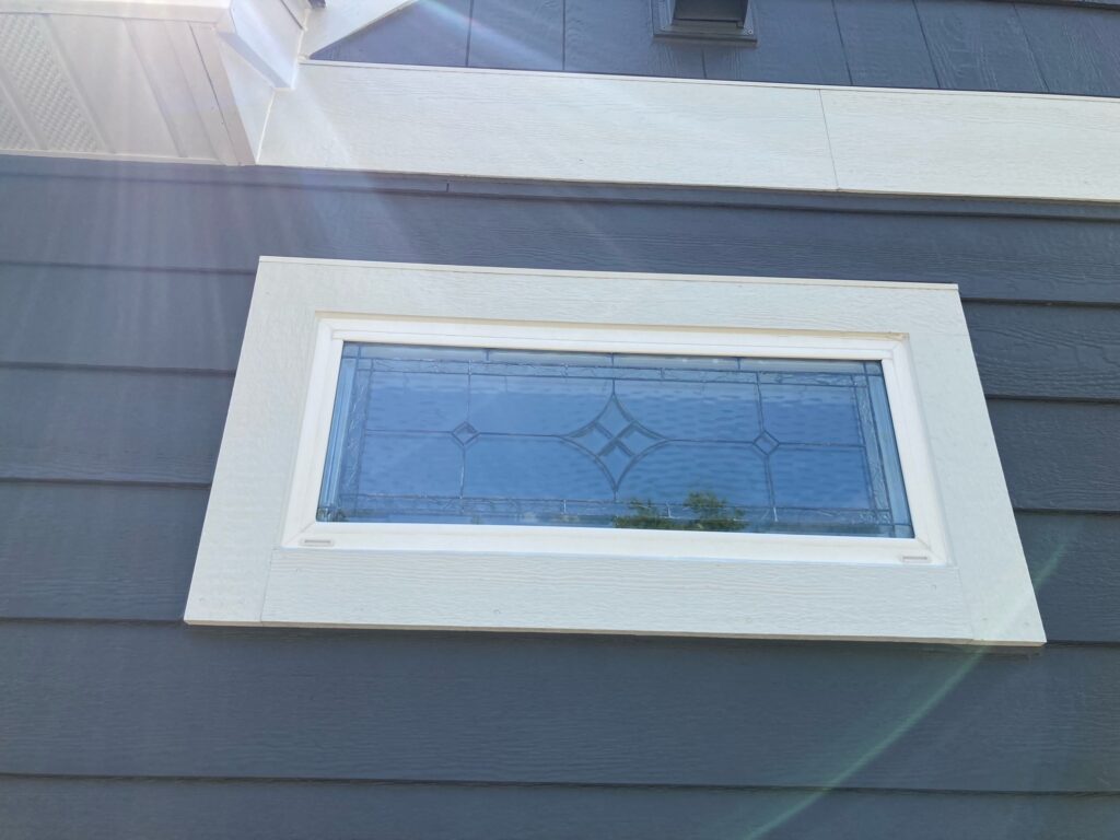 A rectangular exterior window featuring a clear, textured, and leaded stained glass panel with a classic geometric design, including prominent beveled diamond and square accents. The panel is set within a white frame and thick white trim, installed in dark gray horizontal house siding under a white soffit. Sunlight gently reflects off the glass surface, revealing a faint reflection of trees.