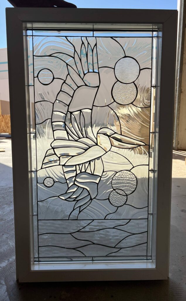 A rectangular stained glass panel in a white frame, featuring an abstract design of a hummingbird with large, sweeping wings and a long beak, crafted from clear textured and beveled glass, accented by circular textured glass elements, set against a background of wavy clear glass with dark lead lines.