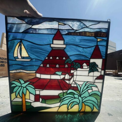 A rectangular stained glass panel, held up against a bright blue sky, depicts a vibrant coastal scene. The central feature is a red-roofed building, reminiscent of the Catalina Casino, surrounded by smaller white structures and lush green palm trees. A sandy beach curves along the bottom, leading into clear blue water where a white sailboat with a golden sail glides. The sky above is a varied blue, suggesting movement, and a distant, darker landmass is visible beyond the water. The panel's lead lines are dark, contrasting with the colorful art glass.