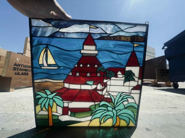 A rectangular stained glass panel, held up against a bright blue sky, depicts a vibrant coastal scene. The central feature is a red-roofed building, reminiscent of the Catalina Casino, surrounded by smaller white structures and lush green palm trees. A sandy beach curves along the bottom, leading into clear blue water where a white sailboat with a golden sail glides. The sky above is a varied blue, suggesting movement, and a distant, darker landmass is visible beyond the water. The panel's lead lines are dark, contrasting with the colorful art glass.