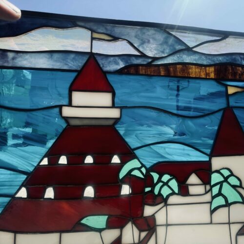 A rectangular leaded stained glass panel depicting a vibrant coastal scene. It features a grand castle with dark red conical roofs, white walls, and small arched white windows, surrounded by green foliage. The textured blue glass forms a lively sea, and light blue and white textured glass represents a cloudy sky. A hand is visible holding the top left edge of the panel.