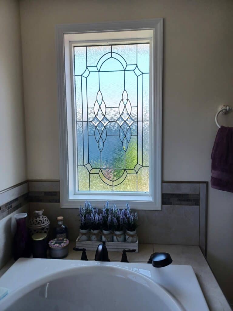 A rectangular clear stained glass window with a central symmetrical geometric pattern featuring textured glass and clear beveled elements, set in a white frame above a bathtub.