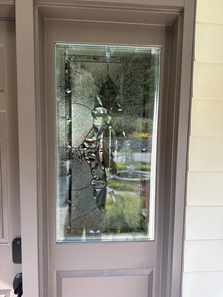 A rectangular clear stained glass door insert featuring a central abstract pattern of polished bevels, surrounded by varied textured privacy glass, set into a taupe-colored door.