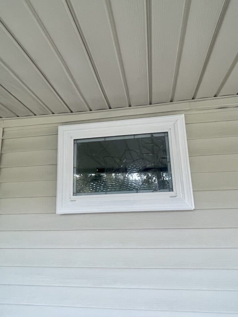 A rectangular clear textured stained glass window in a white frame, installed in light beige horizontal vinyl siding beneath a white soffit. The glass features a clear, abstract pattern of wavy and rippled textures, creating privacy.