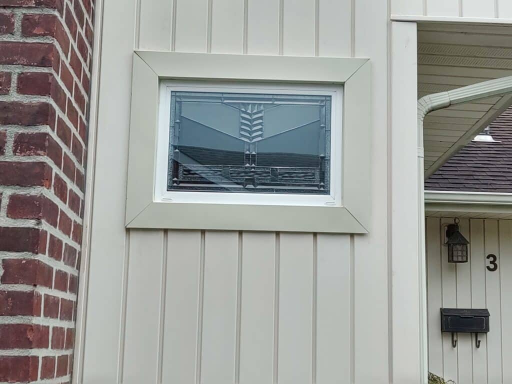 A rectangular clear stained glass insulated window featuring a central stylized geometric design with beveled and textured glass, installed in a light-colored exterior wall.