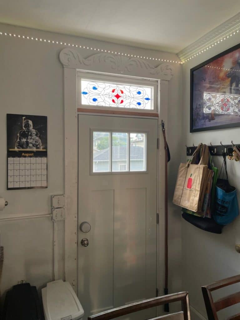A rectangular leaded stained glass transom panel with a clear geometric pattern, featuring a central red diamond and symmetrical blue teardrop shapes, installed above a white interior door.