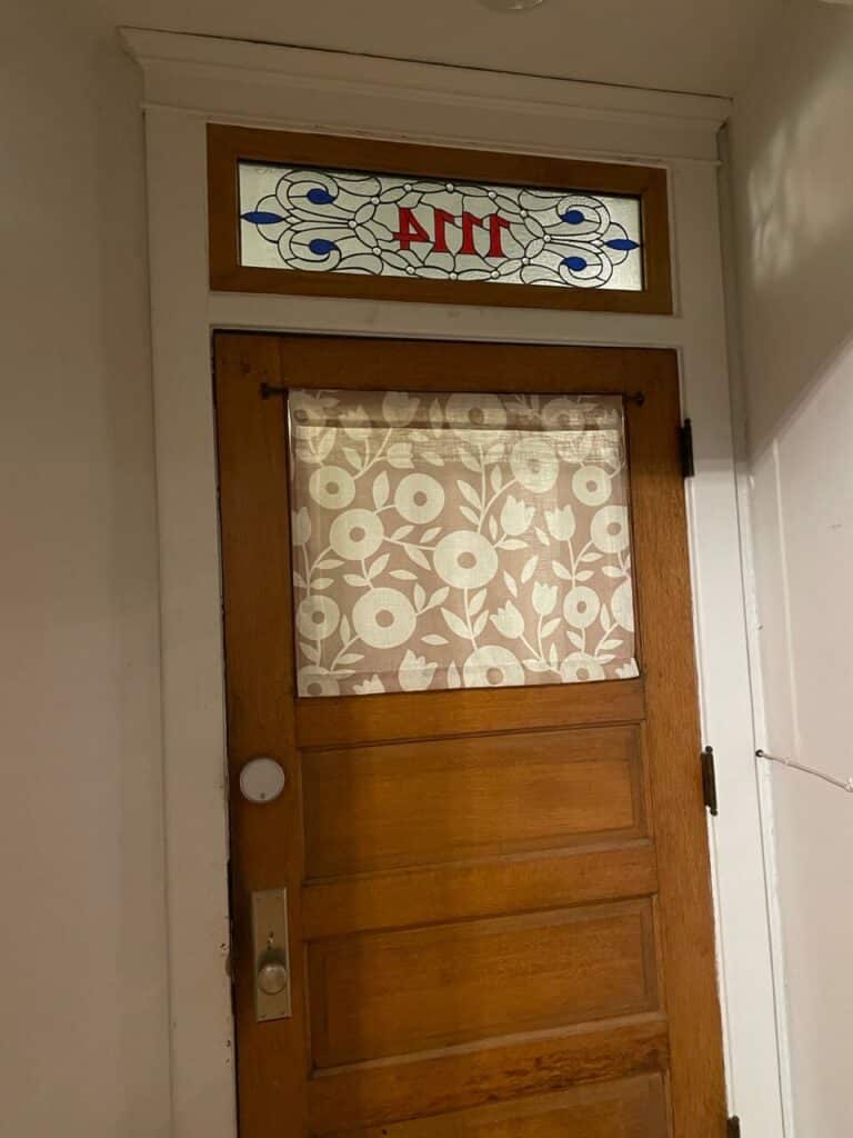 A rectangular stained glass transom window featuring red Greek letters Alpha Pi Pi, surrounded by clear textured glass and blue scrollwork.