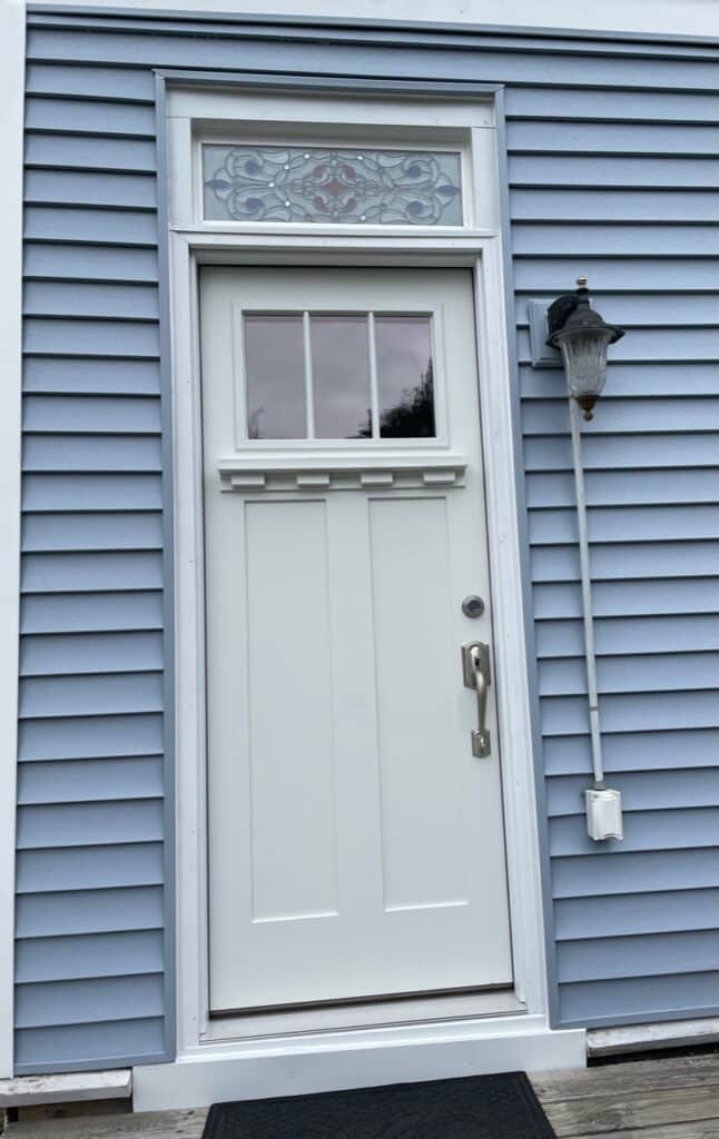A rectangular stained glass transom window featuring an ornate grey, blue, and red scrollwork design, installed above a white front door on a light blue clapboard house.
