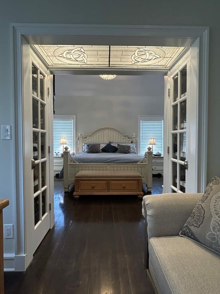 A clear textured stained glass transom panel, featuring two symmetrical Celtic knot designs with lead caming, installed above white French doors leading into a bedroom.