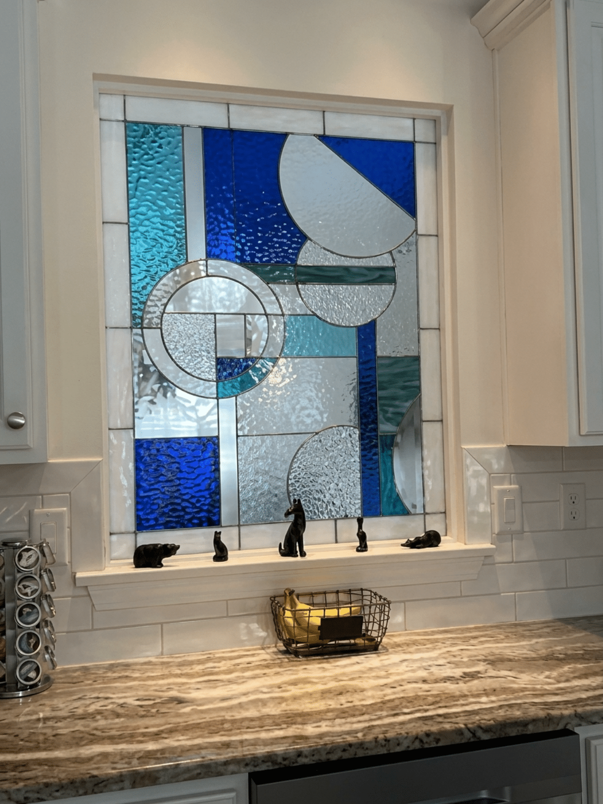 A rectangular, leaded stained glass window featuring an abstract geometric design with intersecting circles, arcs, and rectangular panes in shades of deep blue, aqua, teal, and various clear textured glass.