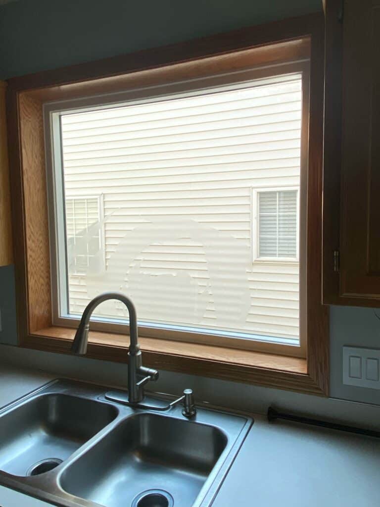 A rectangular window above a kitchen sink, featuring etched glass with irregular, abstract organic shapes for privacy, set in a light wooden frame.