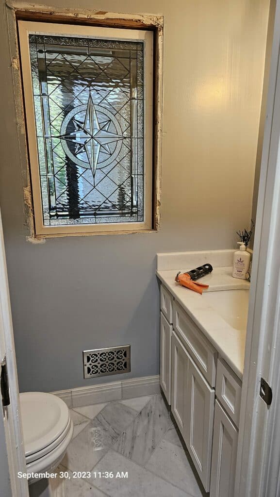 A rectangular, leaded clear stained glass window featuring a central compass rose design with beveled glass. The window is set into a wall above a bathroom vanity, with unfinished trim visible.