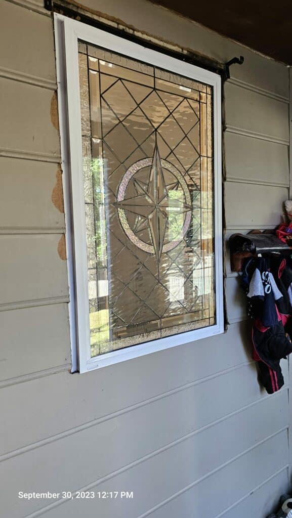 A rectangular, white-framed stained glass window featuring a clear leaded compass rose design with textured and beveled glass, installed in a light-colored exterior wall.